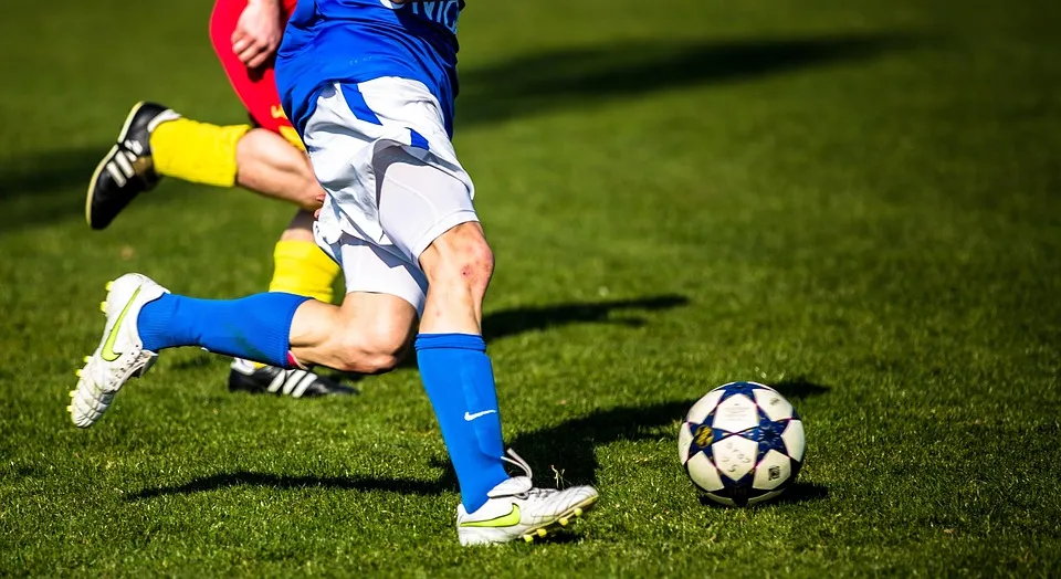 Ein Fußballspieler in blauer Sportkleidung rennt auf einem grünen Spielfeld, während er den Ball mit dem rechten Fuß spielt. Im Hintergrund ist ein anderer Spieler in rot und gelb zu sehen, der sich ebenfalls bewegt.