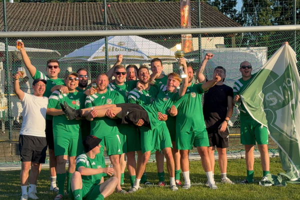 Eine Gruppe von Männern und Frauen in grünen Sporttrikots posiert fröhlich auf einem Fußballfeld. Einige halten eine Flagge, während andere ihre Arme in die Luft heben, um ihren Sieg zu feiern. Im Hintergrund sind Sporteinrichtungen sichtbar.