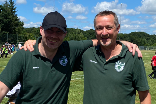 Zwei Männer in grünen Polohemden stehen eng beieinander auf einem Sportfeld, lächelnd und posierend. Im Hintergrund ist ein blauer Himmel zu sehen, gespickt mit wenigen Wolken, sowie grüne Wiesen und Sportaktivitäten.