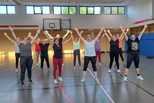 Eine Frau, die in einem Fitnessstudio auf einer roten Matte sitzt und sich nach vorne beugt, um ihr Bein zu dehnen. Sie trägt Sportbekleidung und hat ihren Fokus auf der Dehnung. Im Hintergrund sind Fitnessgeräte sichtbar.