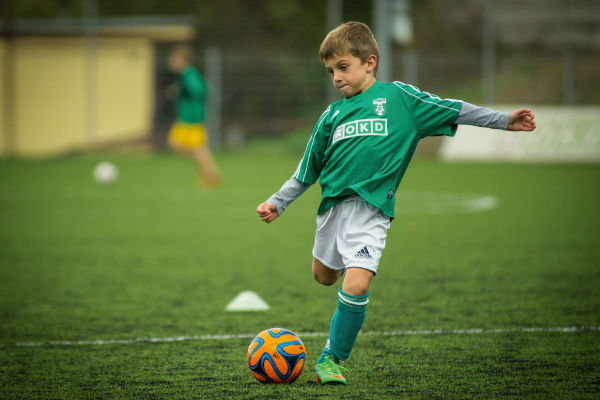 Ein Junge in einem grünen Fußballtrikot tritt einen Fußball auf einem Fußballplatz mit künstlichem Gras. Im Hintergrund sind unscharf weitere Kinder und Spielfeldmarkierungen zu sehen.