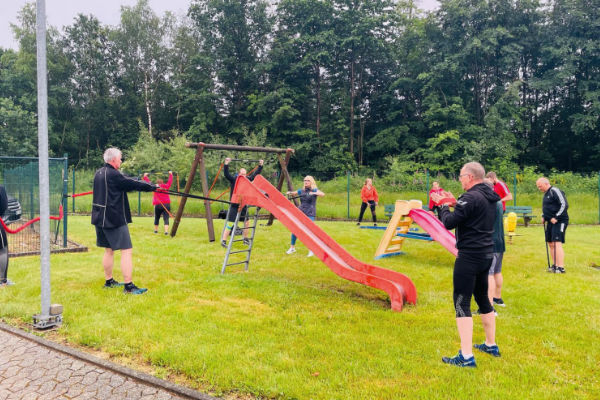 Eine Gruppe von Erwachsenen in Sportkleidung an einer Rutsche auf einem Spielplatz, umgeben von grünen Bäumen. Einige Personen stehen, während andere aktiv sind. Der Platz ist gut gepflegt und es ist bewölkt.