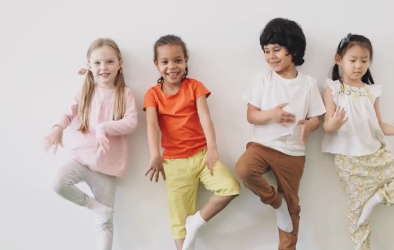 Vier Kinder stehen an einer weißen Wand und machen spielerische Bewegungen. Sie tragen farbenfrohe Kleidung: zwei Mädchen in leichtem Pink und Weiß sowie zwei Jungen in Orange und Braun. Alle lachen und haben eine fröhliche Ausstrahlung.
