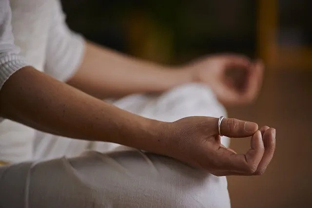 Eine Nahaufnahme einer Hand, die in der Meditationsmudra positioniert ist, während die andere Hand sanft auf einem Oberschenkel ruht. Die Hände sind im Fokus, umgeben von einer beruhigenden, leicht unscharfen Kulisse.